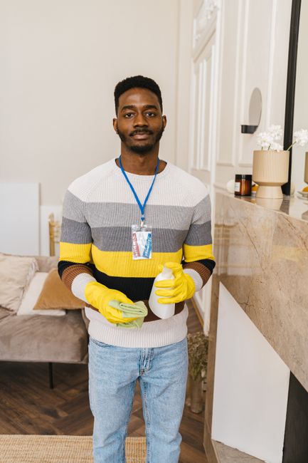A male cleaner standing in a well-lit, modern living room holding cleaning supplies including a spray bottle and a microfiber cloth. He is dressed in casual clothing with yellow rubber gloves and a lanyard around his neck, presenting a professional yet approachable appearance. The background features a beige sofa with cushions, a marble-topped sideboard with decorative vases and a plant, and a bright, tidy space emphasizing cleanliness and hygiene. The scene reflects a domestic environment, illustrating the service of surface cleaning and sanitisation, consistent with the focus of Shepherds Bush Cleaner's affordable domestic cleaning prices for homes in Shepherds Bush.