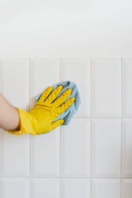 A close-up view of a person wearing a yellow cleaning glove using a blue microfiber cloth to wipe a white tiled wall in a room, likely a kitchen or bathroom. The tiles are glossy and uniformly arranged, showing a clean and shiny surface. The lighting is bright, emphasizing the cleanliness and hygiene of the surface. This image illustrates surface cleaning as part of domestic cleaning services offered by Shepherds Bush Cleaner, demonstrating attention to detail and effective sanitisation of tiled surfaces.