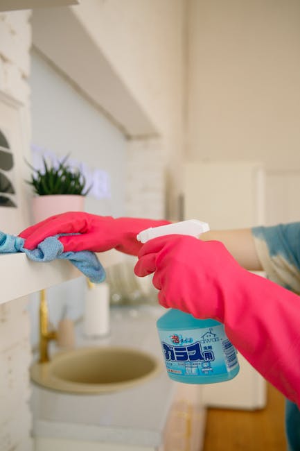 A male cleaner standing in a well-lit, modern living room holding cleaning supplies including a spray bottle and a microfiber cloth. He is dressed in casual clothing with yellow rubber gloves and a lanyard around his neck, presenting a professional yet approachable appearance. The background features a beige sofa with cushions, a marble-topped sideboard with decorative vases and a plant, and a bright, tidy space emphasizing cleanliness and hygiene. The scene reflects a domestic environment, illustrating the service of surface cleaning and sanitisation, consistent with the focus of Shepherds Bush Cleaner's affordable domestic cleaning prices for homes in Shepherds Bush.