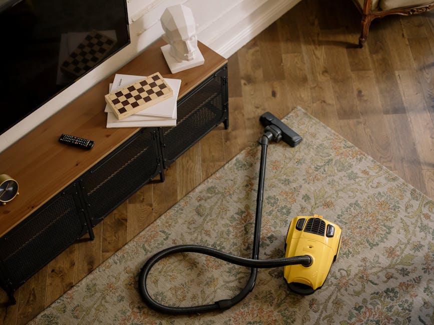 A yellow handheld vacuum cleaner with a flexible hose attachment resting on an ornate, patterned rug in a living room. In the background, a dark wood and metal media unit is positioned against a white wall, topped with a white decorative bust, a stack of white papers and a chess board. A black remote control and a small decorative gold object are also on the media unit. The room features hardwood flooring and is illuminated with natural light, highlighting the clean and tidy appearance of the space. Shepherds Bush Cleaner’s domestic cleaning service is reflected through the presence of the vacuum, indicating surface cleaning and maintenance of the living area.