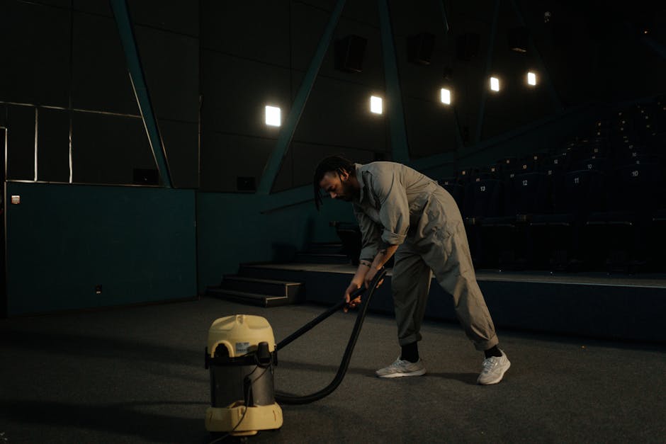 A person operating a yellow and black industrial vacuum cleaner on a dark, carpeted indoor space, possibly a theatre or auditorium, with black tiered seating and blue-green walls in the background. The individual is dressed in light-colored work overalls and is engaged in deep cleaning the carpet surface, with overhead lighting illuminating the scene. The setting appears to be in the process of sanitisation or thorough surface cleaning, as part of professional domestic or commercial cleaning services offered by Shepherds Bush Cleaner near Westfield London Shepherds Bush.