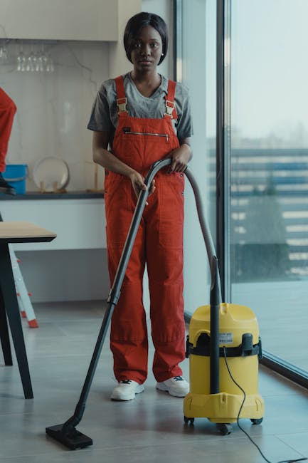 A professional cleaner from Shepherds Bush Cleaner, dressed in a grey t-shirt and red overalls, is operating a yellow commercial vacuum cleaner with a black hose in a modern kitchen. The kitchen features a sleek, light-colored tiled floor that is being deep cleaned, with large glass sliding doors allowing natural light to illuminate the space. Visible surfaces include a light wooden dining table on the left, a white countertop with a glass rack and utensils, and the stainless steel refrigerator in the background. The cleaner stands confidently, showcasing a thorough surface cleaning process for a domestic or rental property end of tenancy, ensuring hygiene and high standards of cleanliness, aligned with Shepherds Bush Cleaner's professional cleaning services.