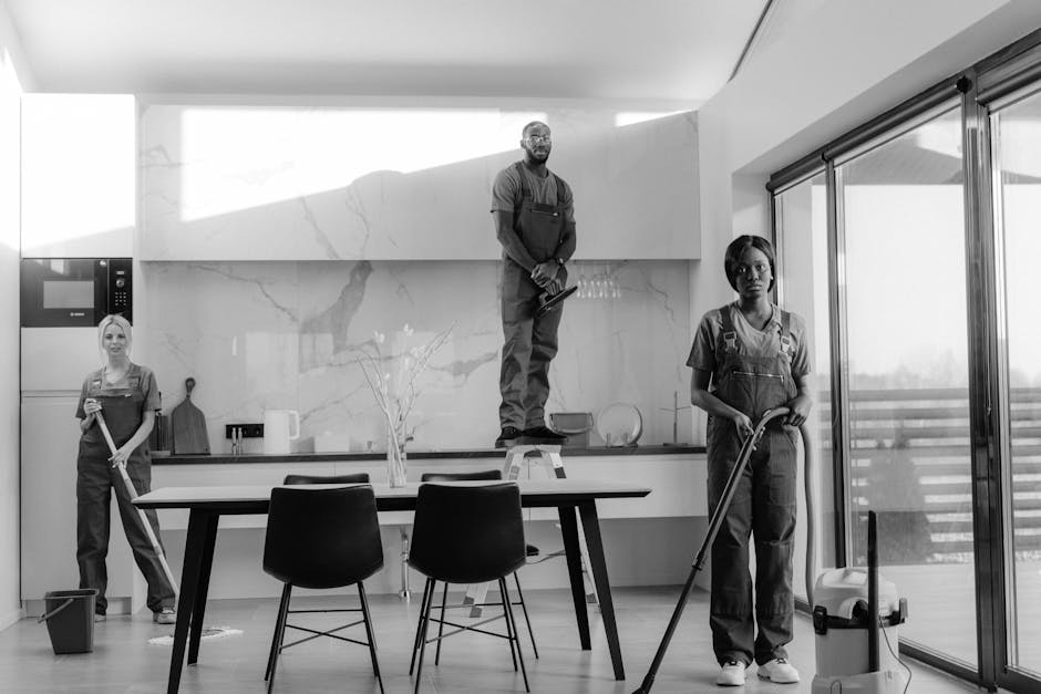 A black and white photograph of three professional cleaners in a modern, bright kitchen. The first cleaner, a woman on the left, stands with a mop and bucket beside her, dressed in overalls. The second cleaner, a man centrally positioned on a step ladder, is using a cleaning cloth on the kitchen upper cabinets, wearing safety glasses and overalls. The third cleaner, a woman on the right, holds a mop and stands next to a vacuum cleaner near large sliding glass doors, which lead to an outdoor balcony with a railing. The kitchen features a smooth marble backsplash, wooden and ceramic surfaces, and a minimalist layout with a dining table and chairs, all illuminated by natural light entering through the glass doors. Shepherds Bush Cleaner’s logo and the clean, hygienic atmosphere emphasize professional surface cleaning and deep cleaning services.