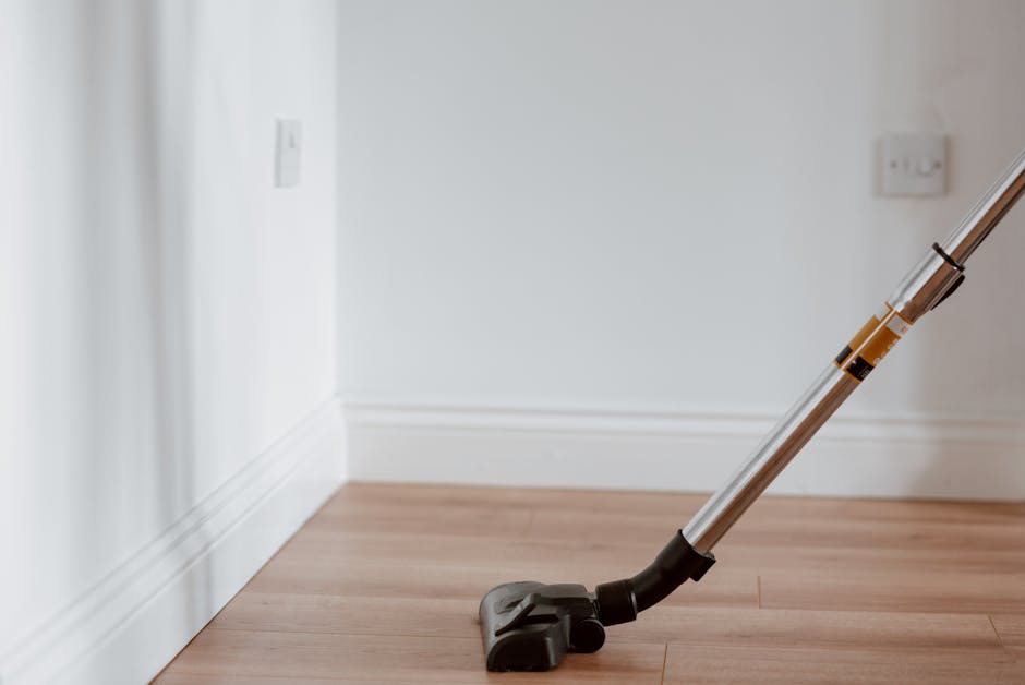 A professional cleaner from Shepherds Bush Cleaner, dressed in a grey t-shirt and red overalls, is operating a yellow commercial vacuum cleaner with a black hose in a modern kitchen. The kitchen features a sleek, light-colored tiled floor that is being deep cleaned, with large glass sliding doors allowing natural light to illuminate the space. Visible surfaces include a light wooden dining table on the left, a white countertop with a glass rack and utensils, and the stainless steel refrigerator in the background. The cleaner stands confidently, showcasing a thorough surface cleaning process for a domestic or rental property end of tenancy, ensuring hygiene and high standards of cleanliness, aligned with Shepherds Bush Cleaner's professional cleaning services.